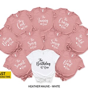 May include: Ten heather mauve t-shirts with white text, each with a different title, such as "The Bossy One", "The Cool One", "The Funny One", "The Sexy One", "The Sassy One", "The Crazy One", "The Smart One", "The Birthday One", "The Bad One", and "The Wild One". One white t-shirt with black text that says "The Birthday One".