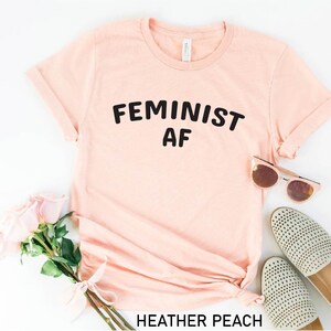 May include: A light pink t-shirt with the text "FEMINIST AF" printed in black. The shirt is laid flat on a white surface with a pair of pink sunglasses and a pair of white shoes.