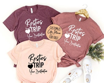 May include: Three t-shirts with the text "Besties Trip" and a heart. The shirts are in maroon, mauve, and peach colours. The text on the shirts is customisable with a city or place.