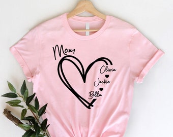 May include: Pink t-shirt with a black heart design and the word "Mom" above it. Inside the heart are the names "Olivia", "Jackie", and "Bella" with a small heart symbol next to each name.