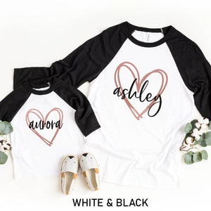 May include: Two white and black raglan shirts with a heart design. One shirt has the name "Aurora" and the other has "Ashley" in a handwritten font. The shirts are displayed on a white surface with decorative elements.