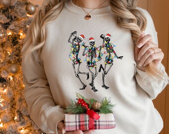 May include: A white sweatshirt with a festive design of three skeletons wearing Santa hats and wrapped in Christmas lights. The skeletons are dancing and smiling.
