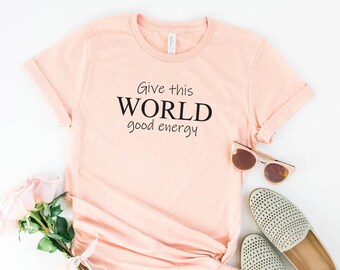 May include: A heather peach colored t-shirt with the text "Give this WORLD good energy" printed on the front.