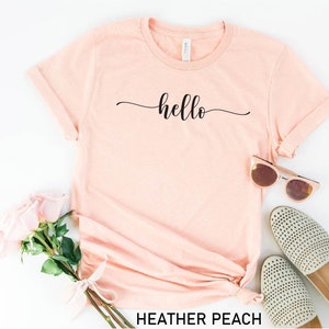 May include: A heather peach colored t-shirt with the word "hello" written in a cursive font on the front.