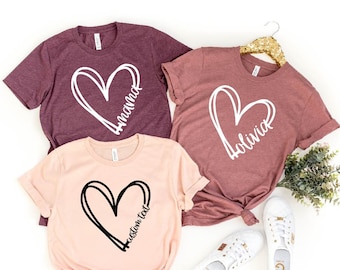 May include: Three t-shirts with a heart design and text. The shirts are in maroon, mauve, and peach colors. The text on the shirts reads "Mama", "Bolivia", and "Custom Text".