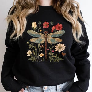 May include: Black sweatshirt featuring a detailed illustration of a dragonfly with blue and gold wings, surrounded by colorful flowers. The design includes red, yellow, and white blossoms, creating a nature-inspired theme.