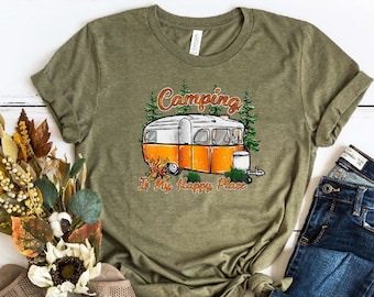 May include: A green t-shirt with a camper van graphic and the text "Camping is my happy place". The shirt is laid out on a white wooden surface with a pair of brown sandals, a bouquet of dried flowers, and a pair of blue jeans.