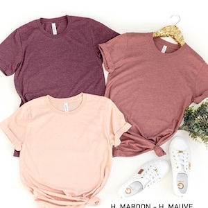 May include: Three plain t-shirts in different colors: maroon, mauve, and peach. The shirts are laid out on a white surface with a sprig of greenery and white sneakers.
