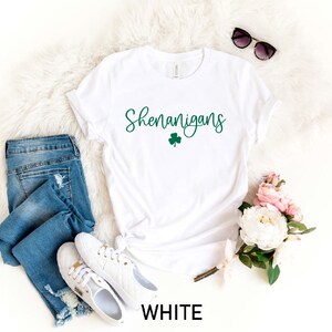 May include: White t-shirt with the text "Shenanigans" in green script with a green shamrock underneath. The shirt is laid out on a white fuzzy surface with a pair of sunglasses, a pair of blue jeans, and a bouquet of pink and white flowers.