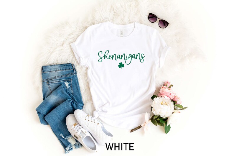 May include: White t-shirt with the text "Shenanigans" in green script with a green shamrock underneath. The shirt is laid out on a white fuzzy surface with a pair of sunglasses, a pair of blue jeans, and a bouquet of pink and white flowers.