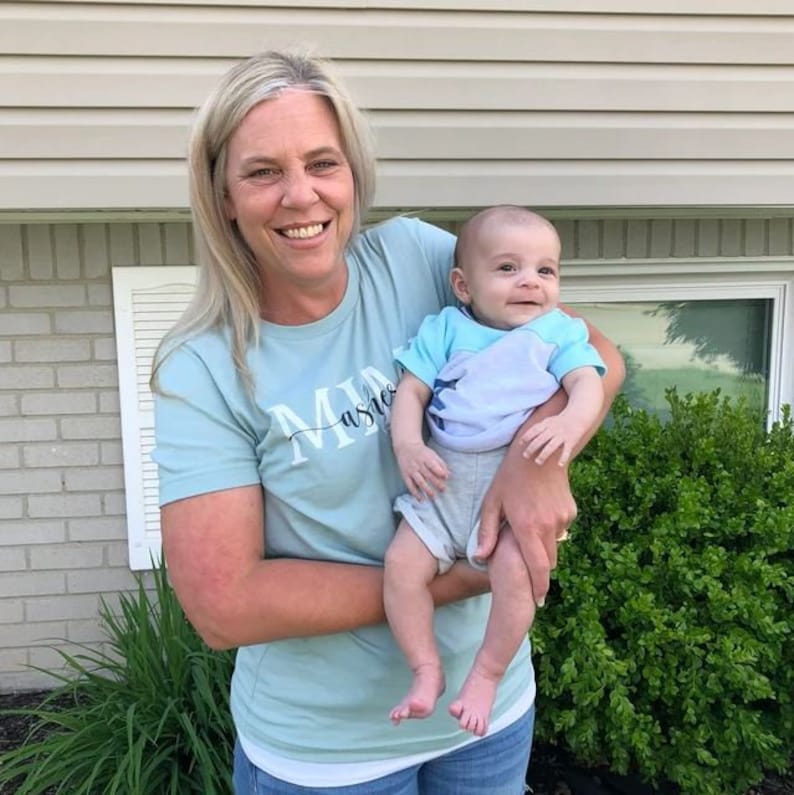 May include: A woman wearing a light blue t-shirt with the text "Mia" on it is holding a baby boy. The baby is wearing a light blue and white striped shirt and gray shorts. The woman and baby are smiling at the camera.