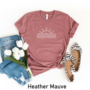 May include: A heather mauve t-shirt featuring a white graphic of a sun setting over waves. The t-shirt is styled with blue jeans and striped bow flats.
