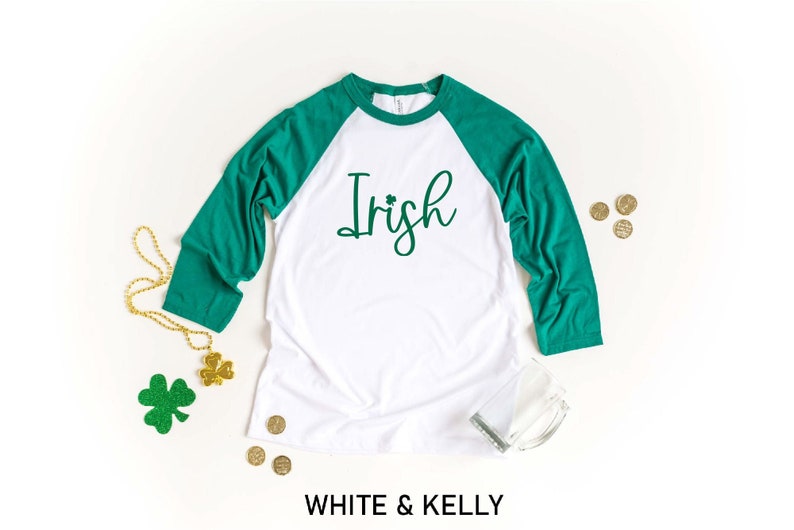 May include: White and green baseball tee shirt with the word "Irish" in green script with a shamrock. The shirt is on a white background with gold shamrocks and beads.