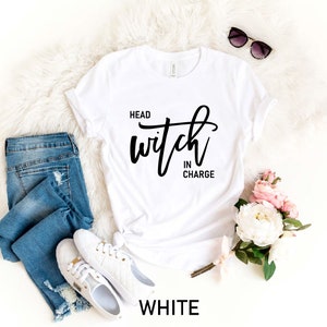 May include: White t-shirt with the text "Head Witch in Charge" in black script lettering. The shirt is laid flat on a white fuzzy surface with a pair of sunglasses, a pair of jeans, and a bouquet of pink and white flowers.