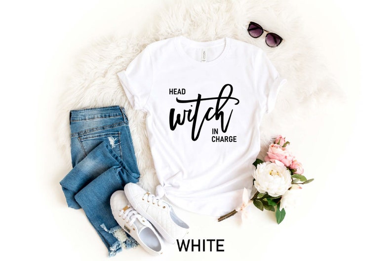 May include: White t-shirt with the text "Head Witch in Charge" in black script lettering. The shirt is laid flat on a white fuzzy surface with a pair of sunglasses, a pair of jeans, and a bouquet of pink and white flowers.