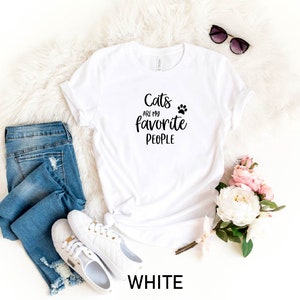 May include: White t-shirt with the text "Cats are my favorite people" and a paw print. The shirt is laying on a white fluffy surface with a pair of jeans, white sneakers, sunglasses, and pink flowers.