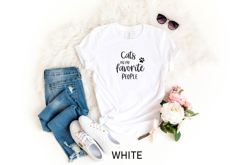 May include: White t-shirt with the text "Cats are my favorite people" and a paw print. The shirt is laying on a white fluffy surface with a pair of jeans, white sneakers, sunglasses, and pink flowers.