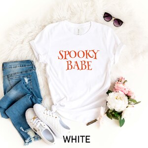 May include: White t-shirt with the text "Spooky Babe" in orange lettering. The shirt is laid flat on a white fuzzy surface with a pair of sunglasses, a pair of blue jeans, and a bouquet of white and pink flowers.