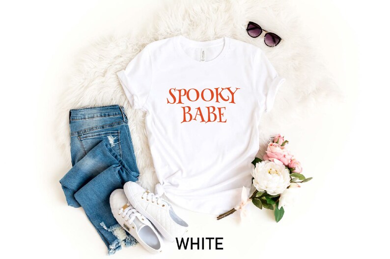 May include: White t-shirt with the text "Spooky Babe" in orange lettering. The shirt is laid flat on a white fuzzy surface with a pair of sunglasses, a pair of blue jeans, and a bouquet of white and pink flowers.