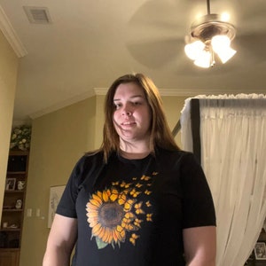 K&ouml;nnte beinhalten: Ein schwarzes T-Shirt mit einem Sonnenblumen-Motiv und gelben Schmetterlingen, die von der Sonnenblume wegfliegen.