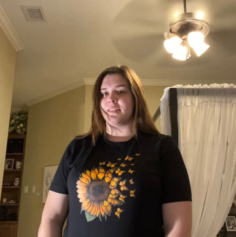 K&ouml;nnte beinhalten: Ein schwarzes T-Shirt mit einem Sonnenblumen-Motiv und gelben Schmetterlingen, die von der Sonnenblume wegfliegen.