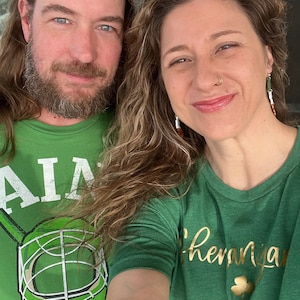 May include: Two people wearing green t-shirts with shamrock designs. The person on the left is wearing a t-shirt with a hockey mask and the text "AIA". The person on the right is wearing a t-shirt with the text "Shenanigans" and "WAREN".