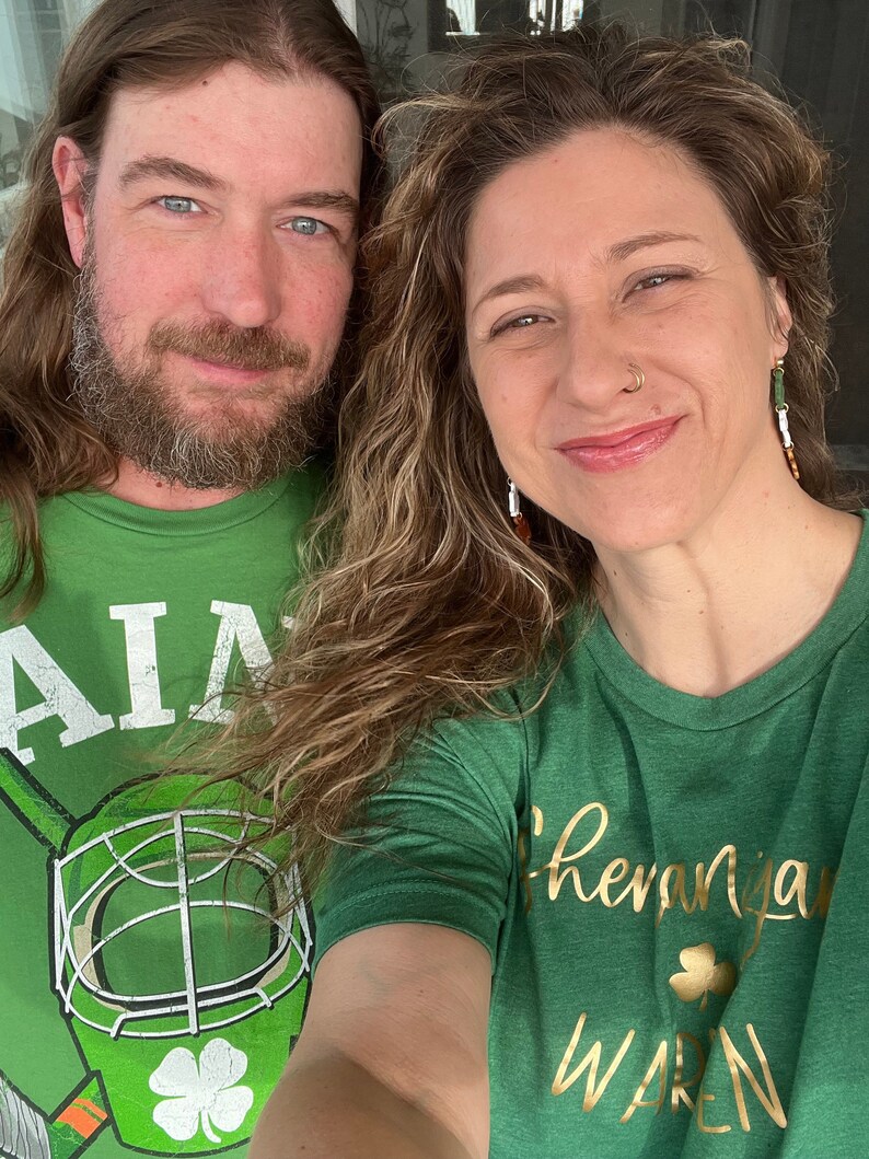 May include: Two people wearing green t-shirts with shamrock designs. The person on the left is wearing a t-shirt with a hockey mask and the text "AIA". The person on the right is wearing a t-shirt with the text "Shenanigans" and "WAREN".