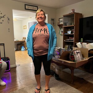 May include: A woman wearing a blue hoodie over a pink t-shirt with the text "Mimi" and "Danielle & Savannah" printed on it. She is wearing black leggings and black flip-flops. She is standing in a living room with a wooden floor and a rug.