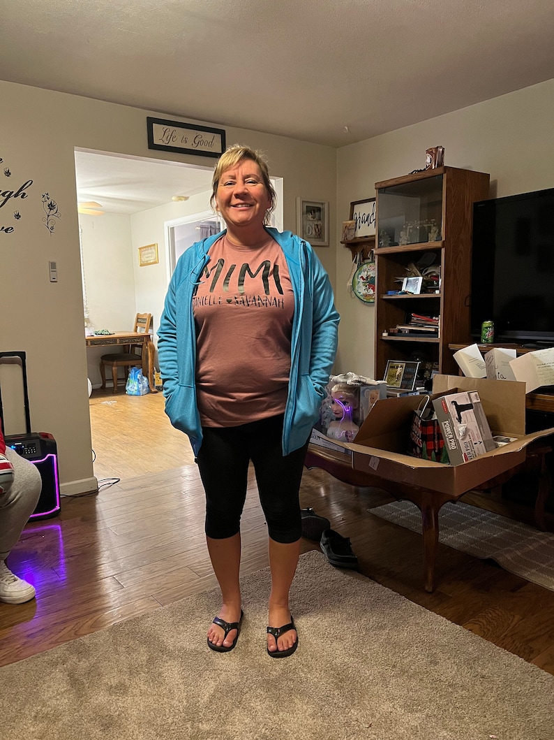 May include: A woman wearing a blue hoodie over a pink t-shirt with the text "Mimi" and "Danielle & Savannah" printed on it. She is wearing black leggings and black flip-flops. She is standing in a living room with a wooden floor and a rug.