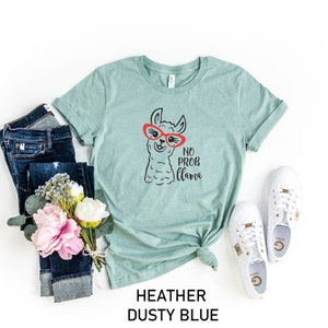May include: A heather dusty blue t-shirt with a black and white illustration of a llama wearing red sunglasses. The text "No Prob Llama" is printed below the llama.
