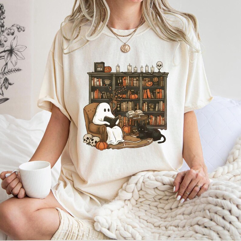 May include: A white t-shirt with a graphic of a ghost reading a book in a chair next to a bookshelf filled with books and a black cat. The bookshelf is decorated with pumpkins and candles.