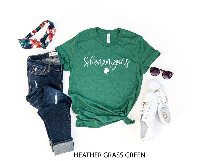 May include: A green t-shirt with the word "Shenanigans" and a shamrock printed in white. The shirt is on a white background with a pair of jeans, a headband, sunglasses, and white sneakers.