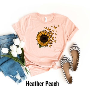 K&ouml;nnte beinhalten: Ein pfirsichfarbenes T-Shirt mit einem Sonnenblumen- und Schmetterlingsmotiv. Das Shirt liegt flach auf einem wei&szlig;en Hintergrund mit einem Paar gestreiften Ballerinas und einer blauen Jeans mit wei&szlig;en Blumen.