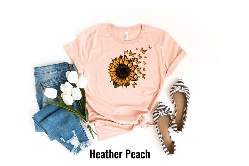 K&ouml;nnte beinhalten: Ein pfirsichfarbenes T-Shirt mit einem Sonnenblumen- und Schmetterlingsmotiv. Das Shirt liegt flach auf einem wei&szlig;en Hintergrund mit einem Paar gestreiften Ballerinas und einer blauen Jeans mit wei&szlig;en Blumen.