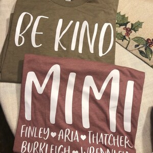 May include: Two t-shirts, one olive green and one maroon, with white text. The olive green shirt says "BE KIND". The maroon shirt says "MIMI" with the names "FINLEY", "ARIA", "THATCHER", "BURKLEY", and "WRENLEY" underneath.