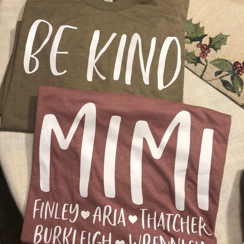 May include: Two t-shirts, one olive green and one maroon, with white text. The olive green shirt says "BE KIND". The maroon shirt says "MIMI" with the names "FINLEY", "ARIA", "THATCHER", "BURKLEY", and "WRENLEY" underneath.