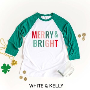 Puede incluir: Camiseta de b&eacute;isbol blanca y verde con el texto "Merry & Bright" en rojo, rosa y verde.