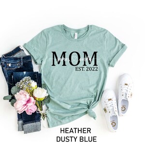 May include: A light blue t-shirt with the text "MOM" in black, with the words "James & Emily" and a heart symbol underneath. The text "EST. 2022" is below the heart symbol.