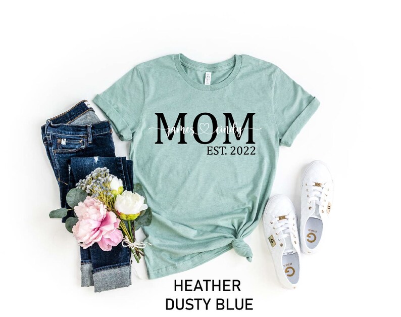 May include: A light blue t-shirt with the text "MOM" in black, with the words "James & Emily" and a heart symbol underneath. The text "EST. 2022" is below the heart symbol.