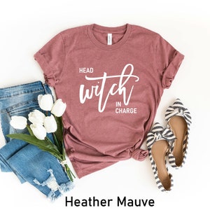 May include: Heather mauve t-shirt with the text "Head Witch in Charge" in white lettering. The shirt is shown with blue jeans and striped flats.