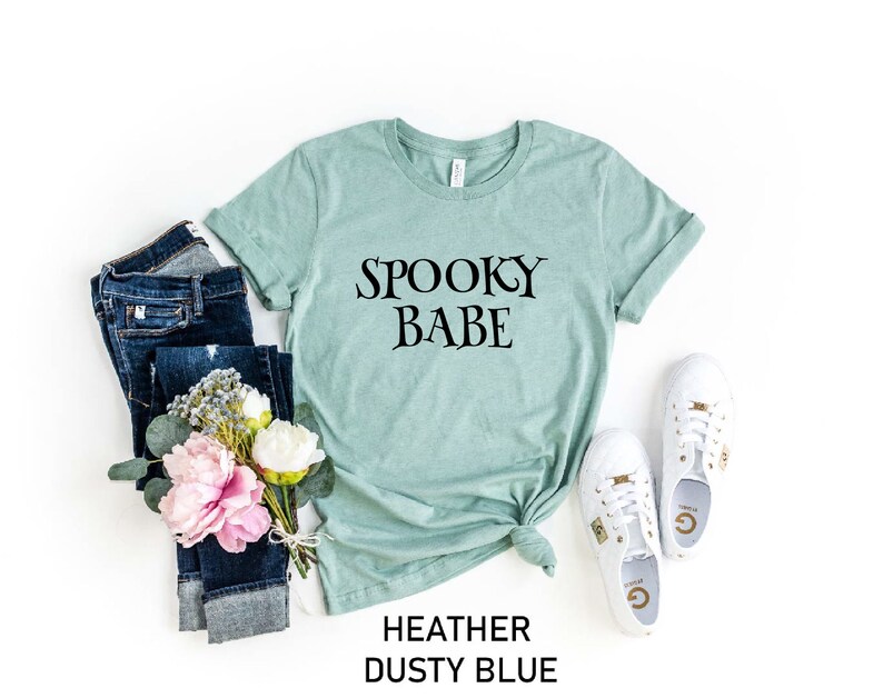 May include: A heather dusty blue t-shirt with the text "Spooky Babe" printed in black. The shirt is styled with blue jeans, a bouquet of pink and white flowers, and white sneakers.