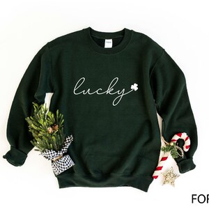 May include: A dark green sweatshirt with the word "lucky" written in white cursive script and a small white shamrock to the right of the word. The sweatshirt is laying flat on a white background.