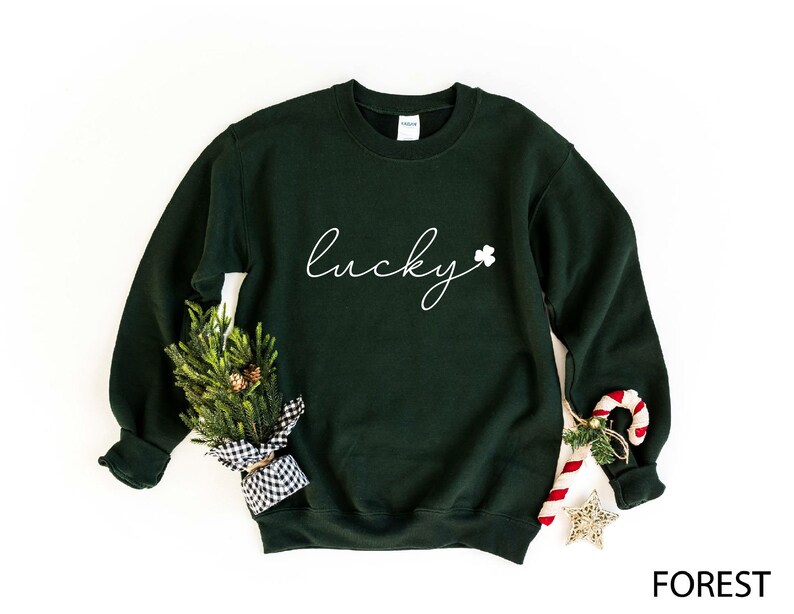 May include: A dark green sweatshirt with the word "lucky" written in white cursive script and a small white shamrock to the right of the word. The sweatshirt is laying flat on a white background.