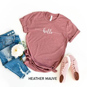 May include: A heather mauve t-shirt with the word "hello" written in white cursive script. The shirt is laid flat on a white background with a pair of pink sneakers and blue jeans.