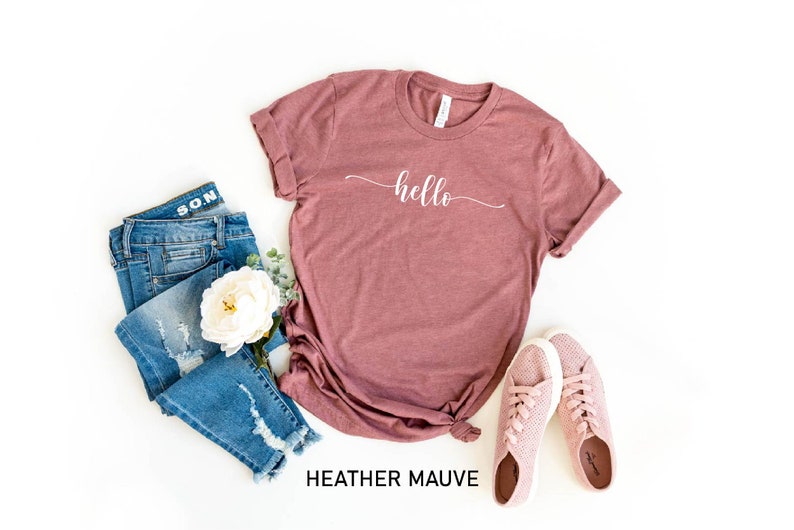 May include: A heather mauve t-shirt with the word "hello" written in white cursive script. The shirt is laid flat on a white background with a pair of pink sneakers and blue jeans.