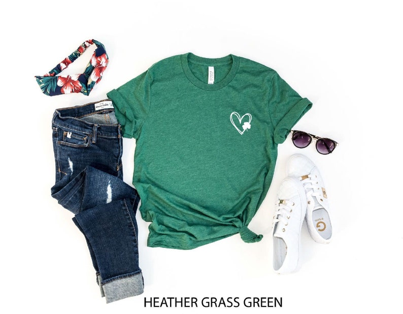 Op de afbeelding: Een groen T-shirt met een wit hart en klavertje vier design. Het shirt wordt gecombineerd met een blauwe jeans, een bloemen hoofdband, witte sneakers en een zonnebril.