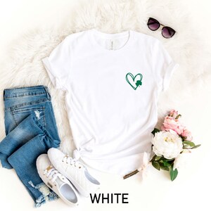 Op de afbeelding: Wit T-shirt met een groene klavertjevier-hart graphic. Het shirt ligt op een wit, pluche oppervlak met blauwe jeans, witte sneakers, een zonnebril en een boeket witte en roze bloemen.
