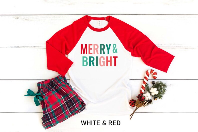 Puede incluir: Camiseta de b&eacute;isbol blanca y roja con el texto "Merry & Bright" en verde, rosa y azul. La camiseta est&aacute; sobre un fondo de madera blanca con pantalones de pijama a cuadros rojos y verdes, un bast&oacute;n de caramelo y una rama de vegetaci&oacute;n.