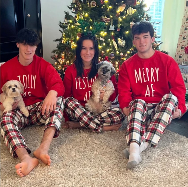 May include: Three people wearing red and white plaid pajamas with festive Christmas-themed text on their shirts. The text on the shirts reads "Jolly", "Festive AF", and "Merry AF". Two small dogs are sitting in front of the people.