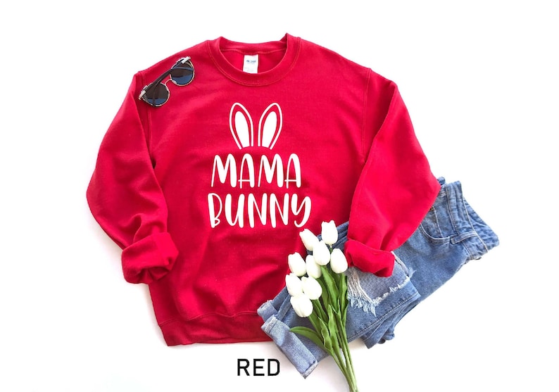 K&ouml;nnte beinhalten: Roter Pullover mit wei&szlig;em Text, der "Mama Bunny" sagt und Hasenohren &uuml;ber dem Text. Der Pullover ist gefaltet und ein Paar Sonnenbrillen und ein Strau&szlig; wei&szlig;er Blumen liegen darauf.
